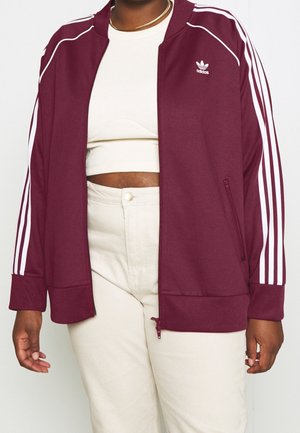 Osoba nosząca bordową kurtkę sportową Adidas z białymi paskami na białym skróconym topie i beżowych spodniach z wysokim stanem, pokazana od szyi do bioder.
