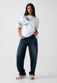 Wit katoenen T-shirt met groene bladborduursel, gecombineerd met donkerblauwe ruime jeans en lichtroze sneakers met zwarte strepen.