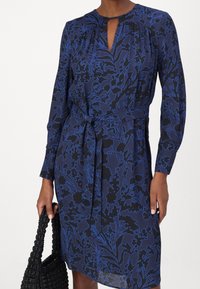 Blommig kjol i marinblått och svart, med blå konturer, långa ärmar, en nyckelhålshalsringning och en knytning i midjan. Bärs med en texturerad svart väska.