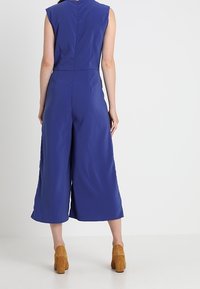 Femme de dos, portant une combinaison sans manches bleue à jambes larges et des bottines marron à talons.