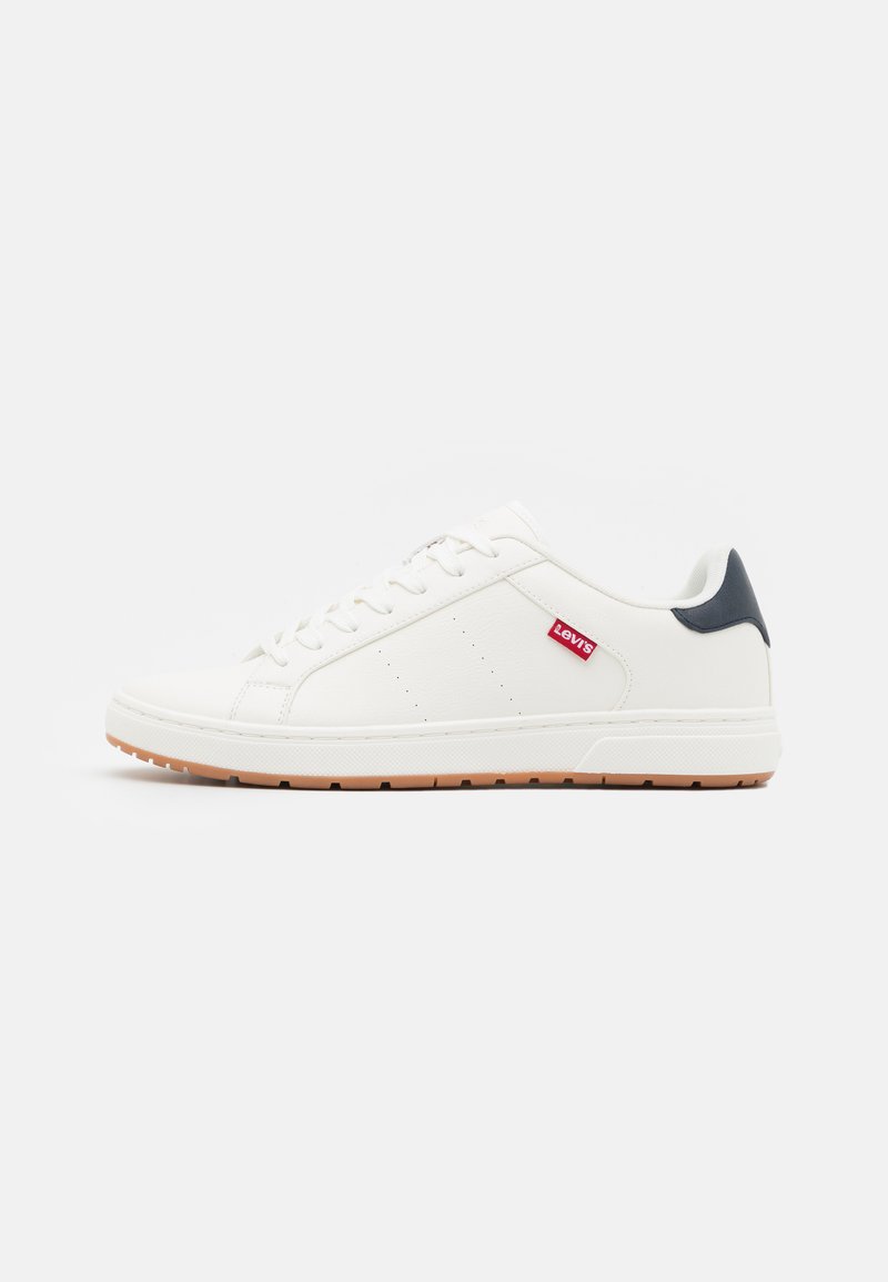 Witte leren sneaker met een gladde afwerking, voorzien van perforaties, een marineblauwe hielaccent en een gumrubberen zool. Rode logo-label aan de zijkant.