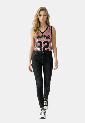 Mujer camina hacia adelante con un top rosa sin mangas de lentejuelas con "92" y vaqueros negros de tiro alto, combinados con zapatillas blancas y negras.