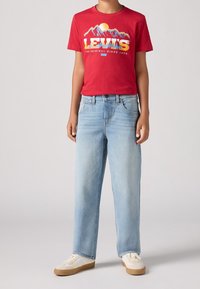 Červené bavlněné tričko s potiskem hor a textem "Levi's", spárované se světle modrými džíny se rovnými nohavicemi a bílými teniskami.