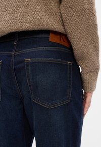 Sötétkék farmernadrág, karcsúsított fazonnal, bőr Calvin Klein címkével a derékrészen és klasszikus hátsó zsebformával.