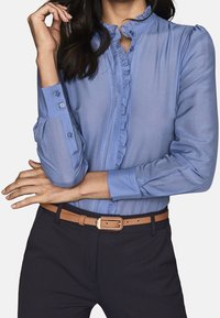 Light blue button-up blouse with ruffled details, long sleeves, and buttoned cuffs. Paired with dark trousers and a brown leather belt.