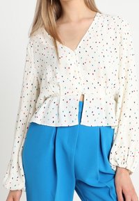 Camicia bianca con pois multicolori, scollo a V, vita arricciata e maniche lunghe a sbuffo. Abbinata a pantaloni blu a vita alta.