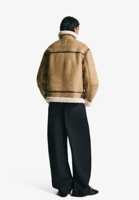 Tan shearlingjacka med vit fleecekrage och bruna läderdetaljer. Kombinerad med svarta vida byxor för en avslappnad passform.