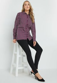 Langszaam, bordeauxrood overhemd met lange mouwen en witte strepen, gecombineerd met zwarte skinny jeans en zwarte hoge hakken, zittend op een witte kruk.