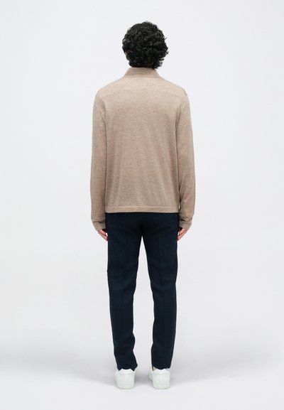 Gai brūns džemperis ar augstu apkakli, garām piedurknēm un taisnu apakšmalu, apvienots ar tumši ziliem biksēm un baltām kedām.