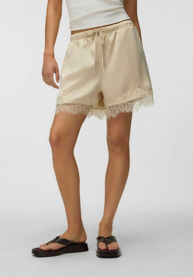 Femme portant un short en satin beige avec bordure en dentelle, un débardeur blanc et des tongs à plateforme noires, debout sur fond blanc.