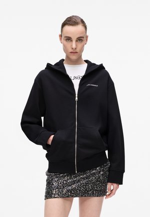 Black zip-up hoodie made of soft fabric, featuring a front pocket and a hood. Worn over a white t-shirt with a grey leopard print. Sparkly mini skirt.