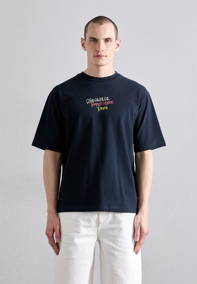 Navy short-sleeve T-shirt in cotton, featuring multicolor embroidered text on the front. Model wears white pants, against a neutral background.