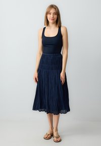 Maglietta senza maniche a coste di colore blu scuro abbinata a una gonna blu scuro plissettata lunga fino al pavimento. La modella indossa sandali beige e si trova davanti a uno sfondo chiaro.