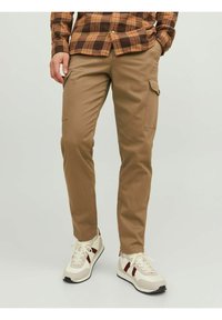 Pantaloni cargo beige con tasche frontali, vestibilità affusolata e texture liscia, abbinati a Sneakers crema e borgogna. Camicia a quadri marrone.