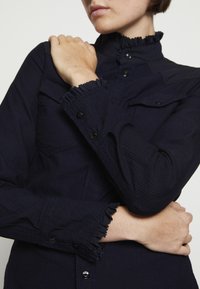 Person klädd i en mörk marinbl å knapp-up skjorta med volangkrage och manschetter, armarna korsade över bröstet, beskuren under ögonen.