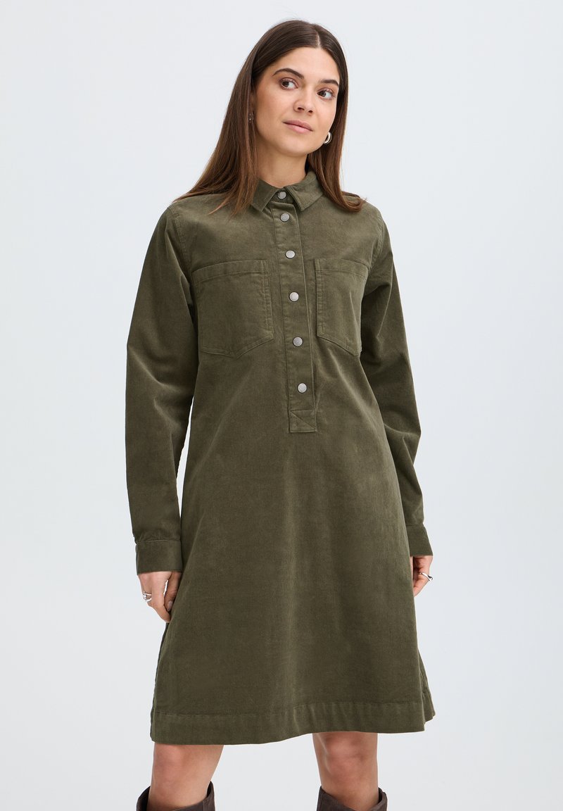 Femme debout regardant sur le côté, portant une robe en velours côtelé vert olive, longueur genou, avec des manches longues et des boutons-pression sur le devant.
