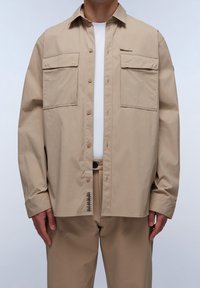Camicia beige con bottoni, realizzata in tessuto leggero, con due tasche sul petto, una pattina frontale e un'etichetta intrecciata sull'orlo.