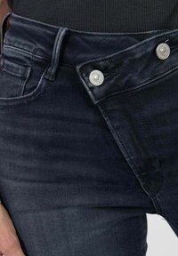Des jeans en denim bleu foncé présentent une taille angulaire unique avec deux boutons-pression en argent et des détails de couture visibles.