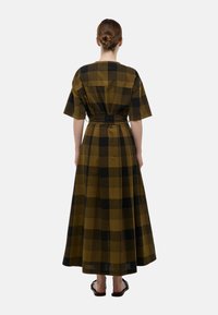 Robe maxi en vert olive et motif à carreaux noirs, manches courtes, taille cintrée, avec une jupe fluide. Texture de tissu lisse, longueur aux chevilles.