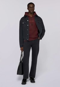 Schwarze Jeansjacke über einem kastanienbraunen Hoodie, kombiniert mit dunkelgrauen Hosen und schwarzen Schuhen. Eine schwarze Tasche haltend, vor hellem Hintergrund stehend.