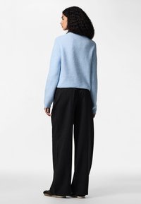 Keeruliste juustega naine, kes kannab helesinist kampsunit, musti laia säärega triibulisi pükse ja musti kingi, seisab valge tausta ees vasakule suunaga.