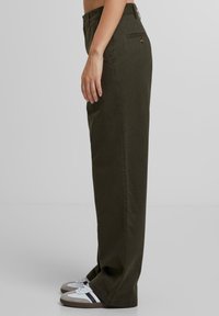 Olijfgroene katoenen broek met een rechte snit, voorzien van een zijzak, een achterste knoop en gedragen met witte en grijze sneakers.