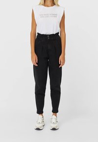 Donna che indossa un top bianco senza maniche con la scritta "Fai sentire il lunedì come se fosse venerdì", pantaloni neri a vita alta e sneakers bianche.