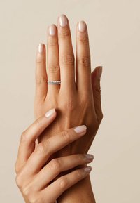 Anello argento con due fasce, una delle quali presenta piccole pietre rotonde. Le mani mostrano una manicure naturale in un colore beige chiaro su uno sfondo neutro.