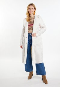 Cappotto lungo color beige chiaro con una texture morbida, bottoni frontali e tasche laterali, indossato sopra un maglione lavorato a maglia multicolore e jeans a gamba larga.