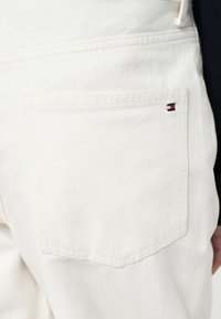 Biele rifľové nohavice s zadným vreckom, na ktorom je malá obdĺžniková červená, biela a modrá značka, ploché švy a jemná textúra.