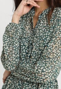 Tummanvihreä kukkakuosinen pusero, jossa kevyt, läpikuultava kangas. Mallissa pieniä valkoisia ja keltaisia kukkia, kauluksellinen pääntie ja kerätyt hihat.