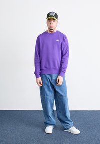 Lilla genser med rund hals og ribbede mansjetter, kombinert med lyseblå baggy jeans. Hvite sneakers fullfører antrekket, sammen med en svart caps med fargerike grafikker.