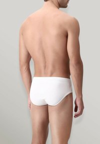 Mutande bianche di cotone che mostrano una vestibilità aderente, una fascia semplice e una texture liscia, viste dal retro su uno sfondo grigio chiaro.