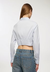 Cameriera blu chiaro a maniche corte con una texture liscia e maniche con bottoni, caratterizzata da una vita arricciata. Indossata con jeans in denim a vita alta.