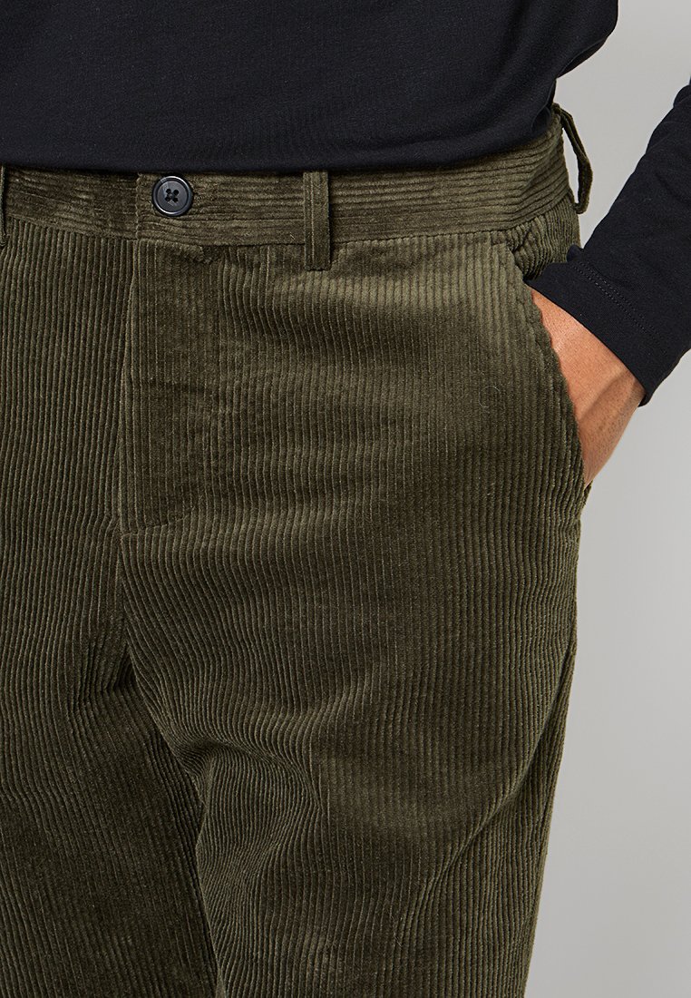 Olivgrüne Cordhosen mit einem strukturierten Rippmuster, ausgestattet mit einer Vordertasche, Gürtelschlaufen und einem einzelnen schwarzen Knopfverschluss.