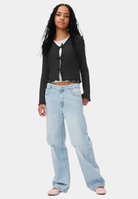 Zwarte geribbelde cropped cardigan met voorbinding, gecombineerd met lichtblauwe losvallende jeans met versleten accenten en roze gevlochten ballerina's.