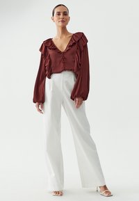 Rust-colored satin blouse with ruffled neckline and sleeves, paired with high-waisted white wide-leg trousers, worn with open-toed heels.