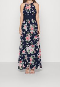 Kvinna klädd i en ärmlös maxiklänning i marinblått med rosa och vita blommönster och en spetsmidja, tillsammans med ljusbeige sandaler med remmar.