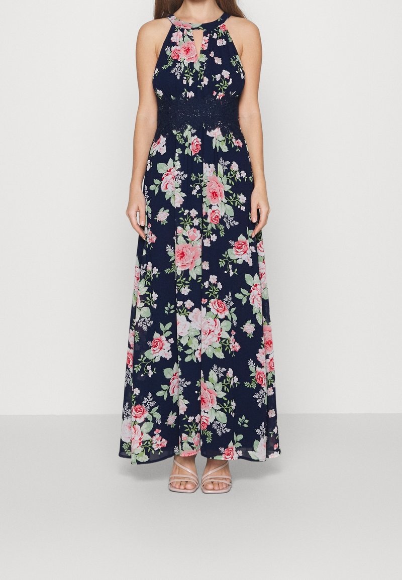 Kvinna klädd i en ärmlös maxiklänning i marinblått med rosa och vita blommönster och en spetsmidja, tillsammans med ljusbeige sandaler med remmar.