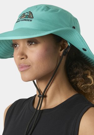 Mujer con un sombrero de ala ancha color verde azulado con correa bajo la barbilla y una blusa sin mangas negra, mirando hacia la izquierda contra un fondo liso.