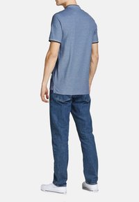 Polo shirt blu a maniche corte con colletto a coste e dettaglio sull'orlo posteriore, abbinato a jeans blu e sneakers bianche. Vestibilità e texture casual.