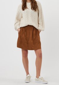 Bruine rok van imitatie-suède met een elastische tailleband en zijzakken, gecombineerd met een crèmekleurige kabelgebreide trui en grijze sneakers.