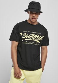 Sort bomulds T-shirt med gul trykt tekst. Kombineret med en sort bucket hat. Modellen har lysegule bukser på og står foran en ensfarvet baggrund.