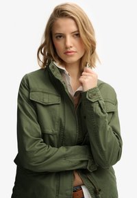 Olijfgroene militaire stijl jas met een rits aan de voorkant, knopenaccenten en twee borstzakken, gemaakt van lichte stof.