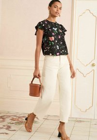 Blusa nera a fiori con maniche arricciate, jeans beige a vita alta, borsa a secchiello marrone e sandali beige con cinturini su pavimento decorato.