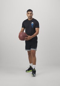 Schwarzes Nike-T-Shirt mit blauer Logo, schwarze Shorts mit weißen Streifen und schwarze Turnschuhe mit grünen Akzenten, einen Basketball haltend.