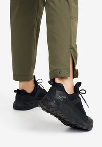 Black athletic shoes with textured soles, featuring a mesh upper and reinforced heel, paired with olive green, tapered trousers.
