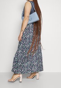 Maxi šaty s tmavě námořnickým podkladem a barevnými vzory ve tvaru kamenů v zelené a růžové barvě. Nosené s světle modrou kabelkou přes rameno a béžovými sandály na bloku.