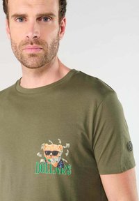 Hombre vistiendo una camiseta verde oliva con un gráfico de un oso con gafas de sol sosteniendo dinero y la palabra "DOLLAR$" en el pecho.