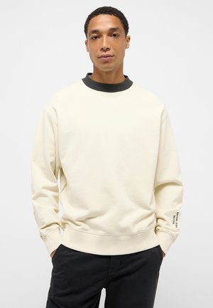 Mann trägt einen cremefarbenen Sweatshirt mit schwarzem Kragen und einem Etikett auf dem linken Ärmel, kombiniert mit schwarzen Hosen, posiert mit den Händen in den Taschen.